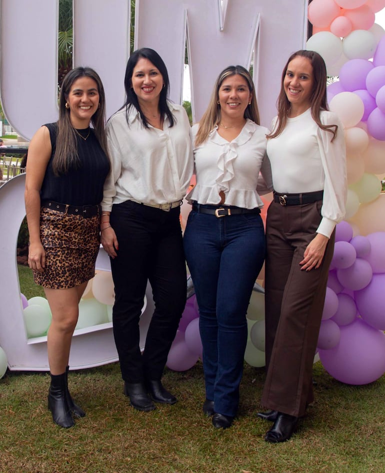 Cuatro mujeres sonríen, vistiendo blusas elegantes blancas y negras, en un entorno festivo decorado con globos de colores suaves.