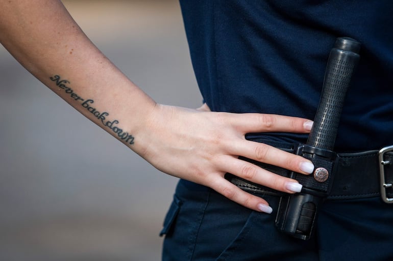 Brazo de oficial de policía con tatuaje que dice 'Never back down', mano en la cintura y porra cercana.