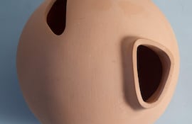 Objeto esférico de cerámica terracota con dos aberturas, presentado en un entorno interior de fondo azul claro.
