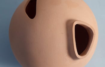 Objeto esférico de cerámica terracota con dos aberturas, presentado en un entorno interior de fondo azul claro.