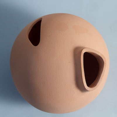 Objeto esférico de cerámica terracota con dos aberturas, presentado en un entorno interior de fondo azul claro.