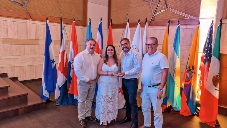 Hombres y mujeres sonrientes en un evento formal; una mujer de vestido blanco recibe un premio de un hombre con camisa blanca.
