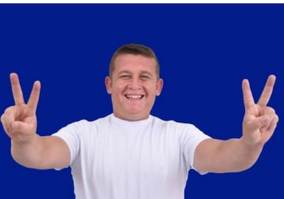 Hombre sonriente con camiseta blanca, mostrando dos dedos en señal de paz, frente a fondo azul liso.