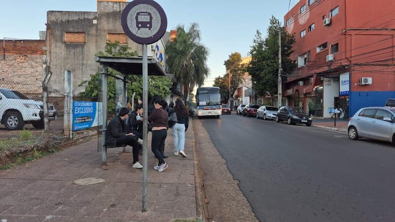 Varias personas esperan en una parada de autobús, algunas sentadas en un banco, en una calle de Encarnación.
