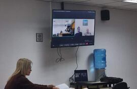 Mujer sentada revisando documentos en una mesa, en ambiente formal y tecnología visible, durante audiencia virtual.