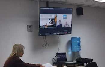 Mujer sentada revisando documentos en una mesa, en ambiente formal y tecnología visible, durante audiencia virtual.