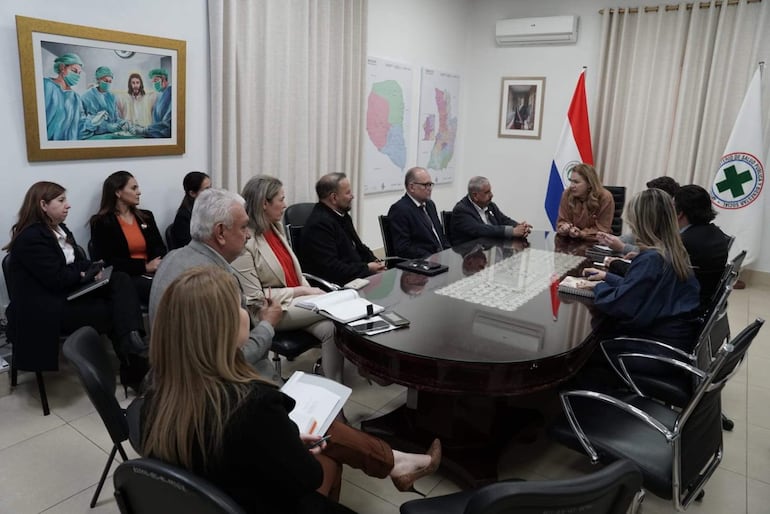 María Teresa Barán, con cabello rubio y largo, habla en reunión con otros asistentes en sala de conferencia, banderas de Paraguay al fondo.
