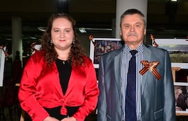 Mujer con blusa roja y falda negra junto a hombre en traje gris, posando en evento con exposiciones fotográficas de fondo.