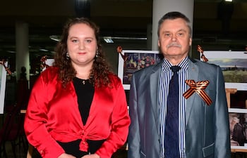 Mujer con blusa roja y falda negra junto a hombre en traje gris, posando en evento con exposiciones fotográficas de fondo.