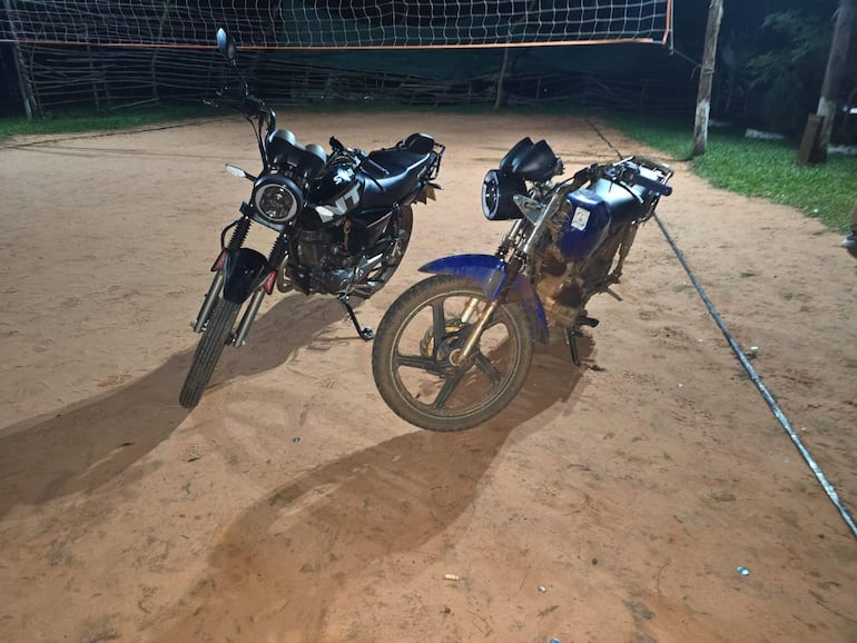 Dos motocicletas en un campo de fútbol iluminado, una negra moderna y una azul en mal estado, sin personas presentes.