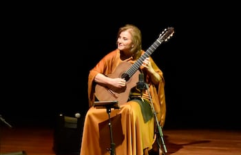 Berta Rojas, con prenda cálida y guitarra, interpreta en un escenario oscuro con luz centrada en ella.