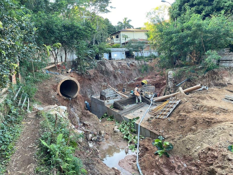Cuatro trabajadores en obra, manipulando tubos y materiales, rodeados de vegetación y escombros en un área de construcción.