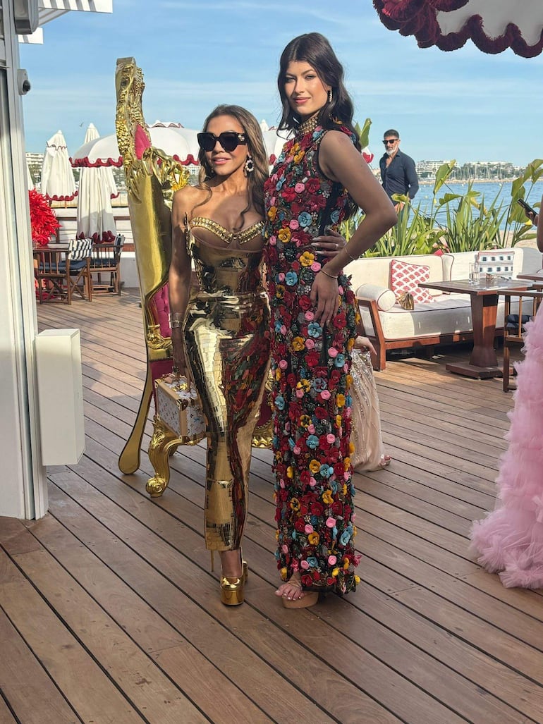 Mujeres sonrientes en vestido dorado y traje floral, rodeadas de un ambiente elegante con vista al mar.