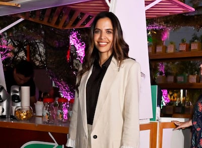 Mujer sonriente en conjunto claro y chaqueta oscura, en ambiente festivo con luces rosas y un hombre detrás de un mostrador.