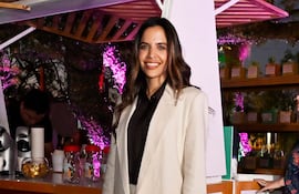 Mujer sonriente en conjunto claro y chaqueta oscura, en ambiente festivo con luces rosas y un hombre detrás de un mostrador.