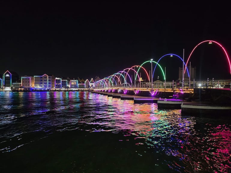 Puente iluminado de varios colores, reflejándose en el agua, con edificios iluminados al fondo en un ambiente nocturno.