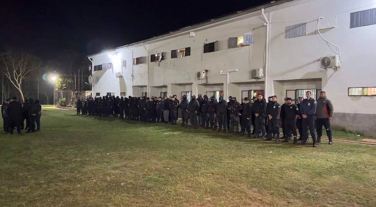 Grupo de 30 a 40 uniformados vestidos de negro en formación en un espacio exterior iluminado, rodeados de árboles.