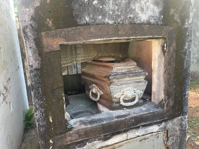 Mausoleo grisáceo con ataúd de madera visible en un cementerio, muestra signos de desgaste y moho.