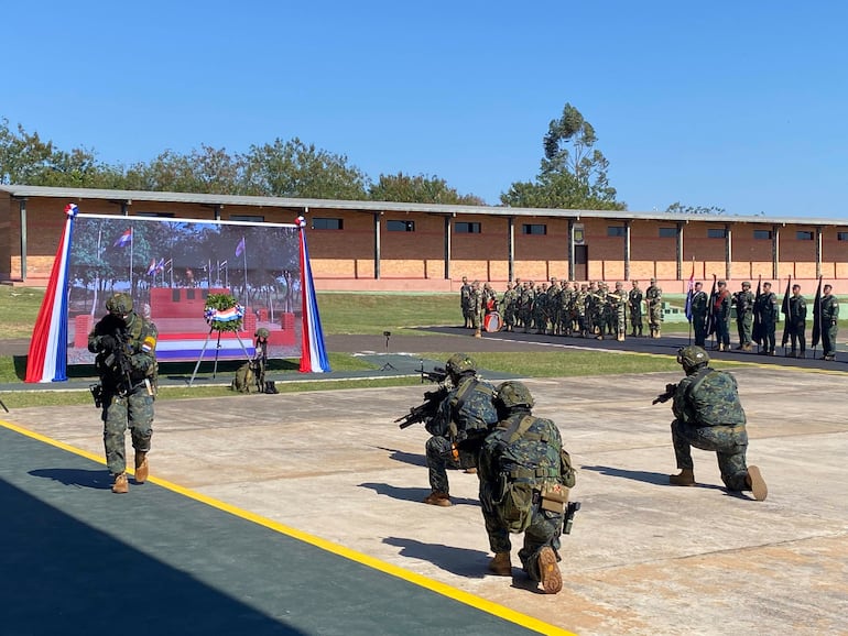 Cuatro soldados en uniforme camuflado, armados, con fondo de pantalla alusiva a la bandera nacional y arreglo floral.