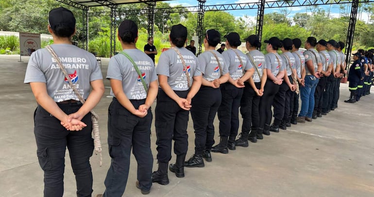 Doce bomberos aspirantes en camisetas grises alineados con manos entrelazadas, bajo una estructura metálica en un entorno floral.