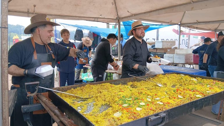 Dos hombres cocinan en carpa, uno con delantal marrón y sombrero claro, el otro con guantes negros. Ambiente festivo y comunitario.