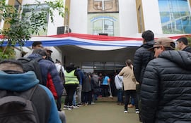 Fila de personas de diferentes edades y géneros, algunas con mochilas, ante un edificio municipal con el escudo visible.