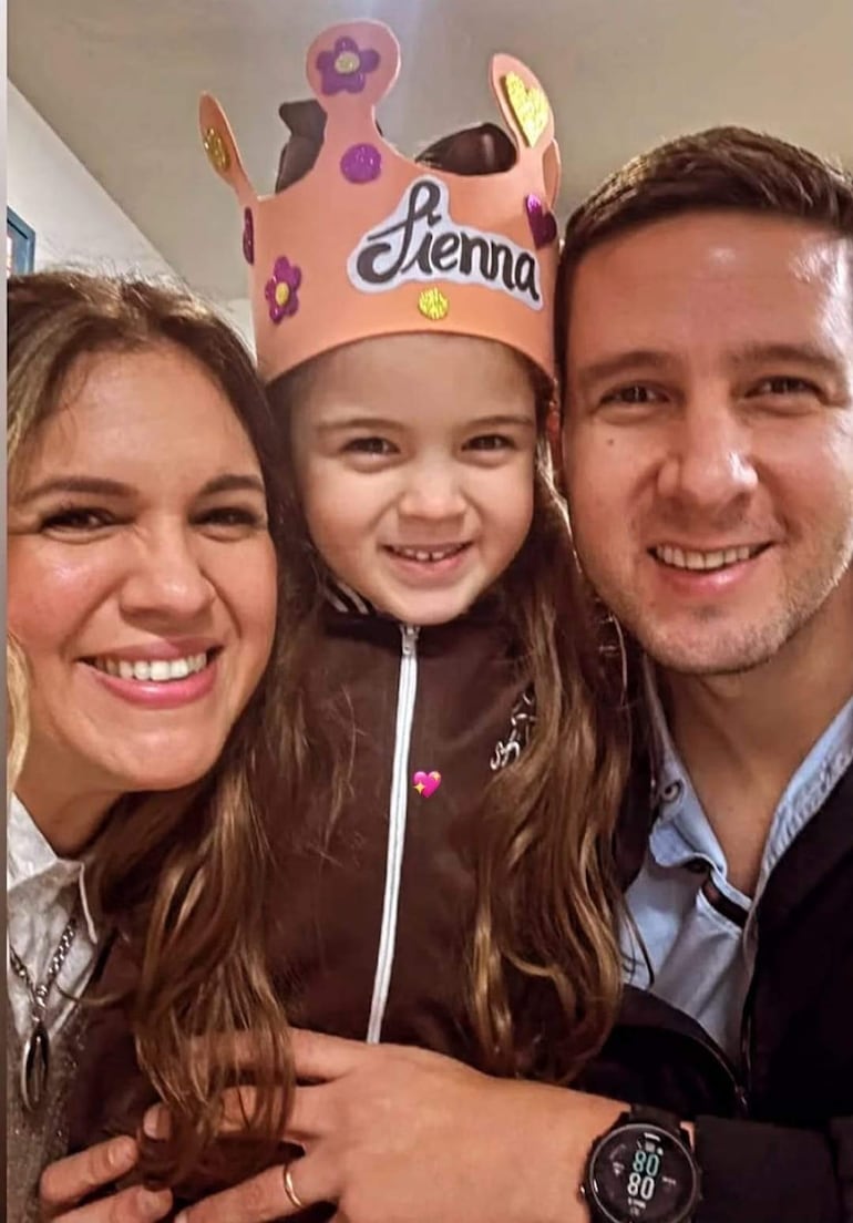 Sienna sonríe con una corona de papel, junto a su madre de cabello rizado y un hombre, todos felices en un ambiente festivo.