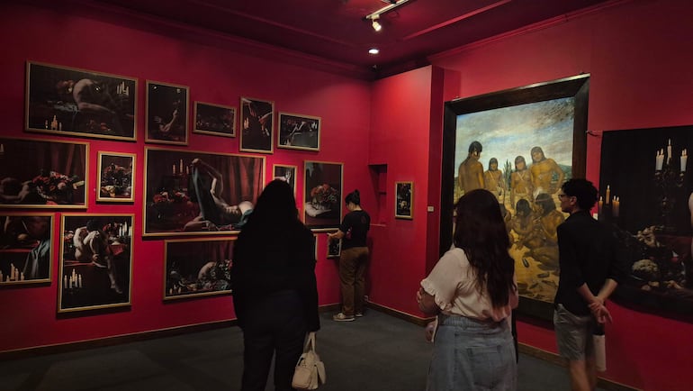 Mujer de pie ajustando obra en galería, vestida de manera casual, rodeada de dos personas que observan las pinturas en muros rojos.