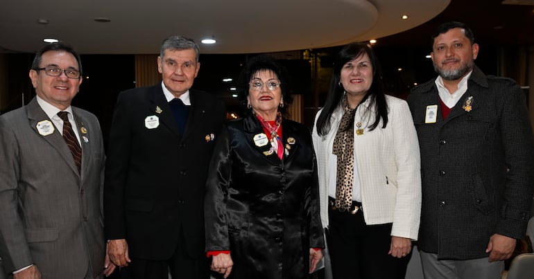 Cinco personas en salón iluminado; mujer central en vestido negro, hombres con chaquetas y mujeres a los lados muestran expresiones felices.