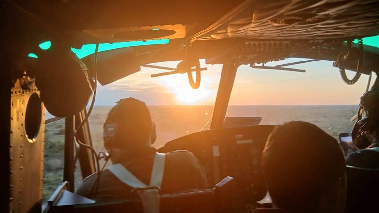 Pilotos con cascos en la cabina de un helicóptero, concentrados en los controles, con paisaje de atardecer visible por el parabrisas.