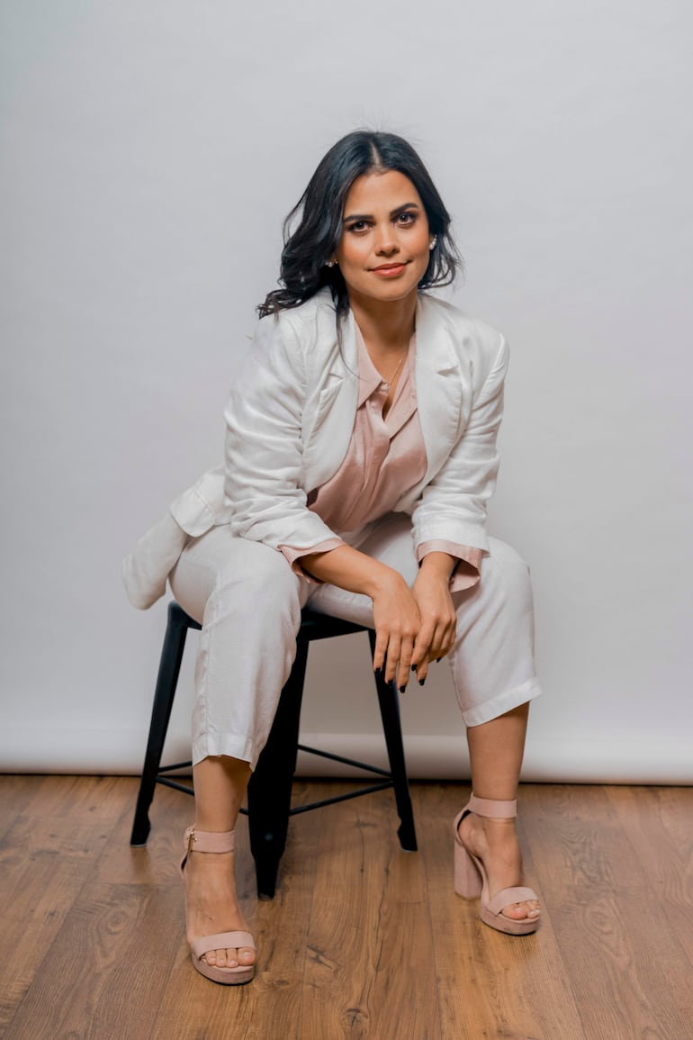 Bianca Parra Roig, mujer de cabello oscuro, vestida con traje claro y blusa rosa, sentada con expresión serena sobre un taburete negro.