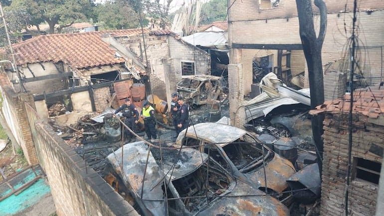 Cuatro personas en uniforme de seguridad conversan en un entorno de incendio, con edificios dañados y vehículos carbonizados de fondo.