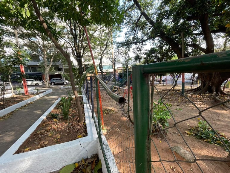 Entorno urbano de parque con sendero pavimentado, árboles y cerca verde. Edificio y vehículos al fondo.