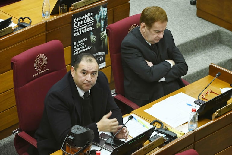 Santiago Amarilla Rios gesticula con seriedad mientras Orlando Penner Durksen escucha con los brazos cruzados en un entorno legislativo.