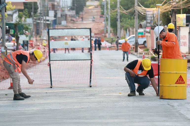 Dos trabajadores, uno agachado y otro de pie en chaleco anaranjado y casco amarillo, miden en una calle de construcción.