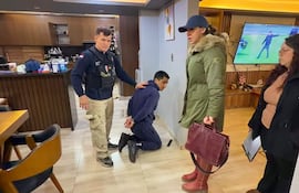 Hombre esposado en el suelo bajo custodia policial, rodeado por dos mujeres y un agente en uniforme oscuro en un ambiente moderno.