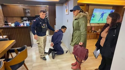 Hombre esposado en el suelo bajo custodia policial, rodeado por dos mujeres y un agente en uniforme oscuro en un ambiente moderno.