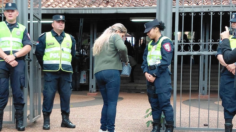 Cuatro oficiales de policía en uniforme negro y verde, mientras una mujer con suéter verde revisa su bolso en un lugar público.