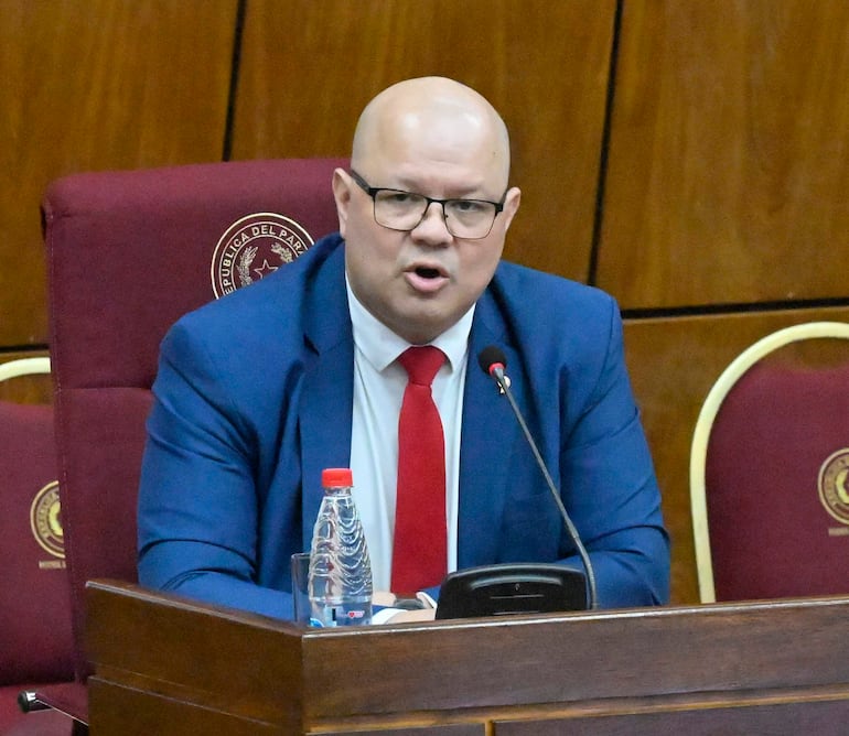 Óscar Lovera, calvo y con gafas, habla desde un estrado con traje azul y corbata roja en un entorno solemne.