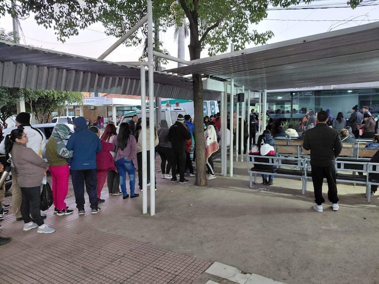 Fila de personas bajo una estructura techada, algunas con abrigos, esperando atención médica en un ambiente urbano.