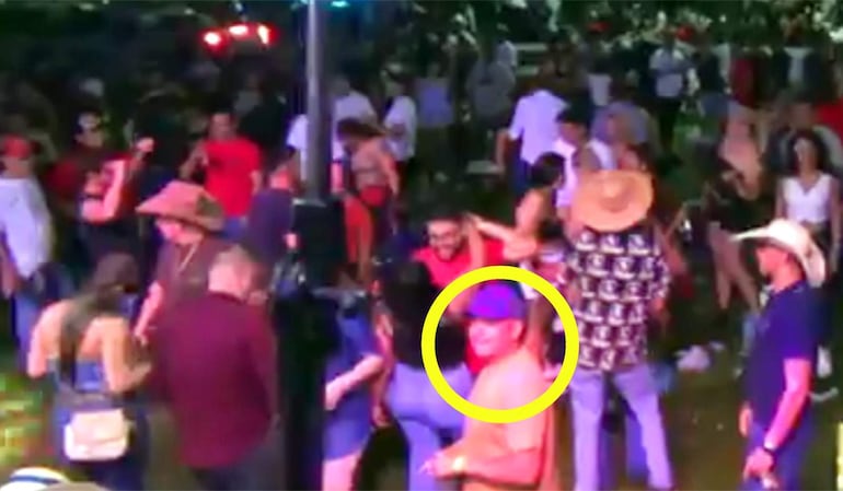 Hombre con camiseta naranja y gorra azul observa a la cámara en un club, rodeado de personas bailando y disfrutando de la fiesta.