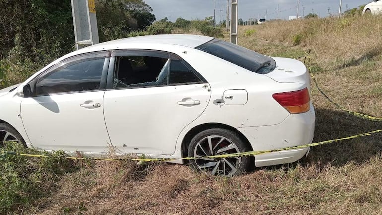 Automóvil blanco estacionado con daños visibles, rodeado de cinta de seguridad amarilla en un área verde descuidada.