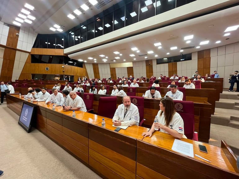 Grupo de varias personas conversando en un ambiente formal, con camisas blancas y sillas burdeos, en una reunión de sociedades médicas.