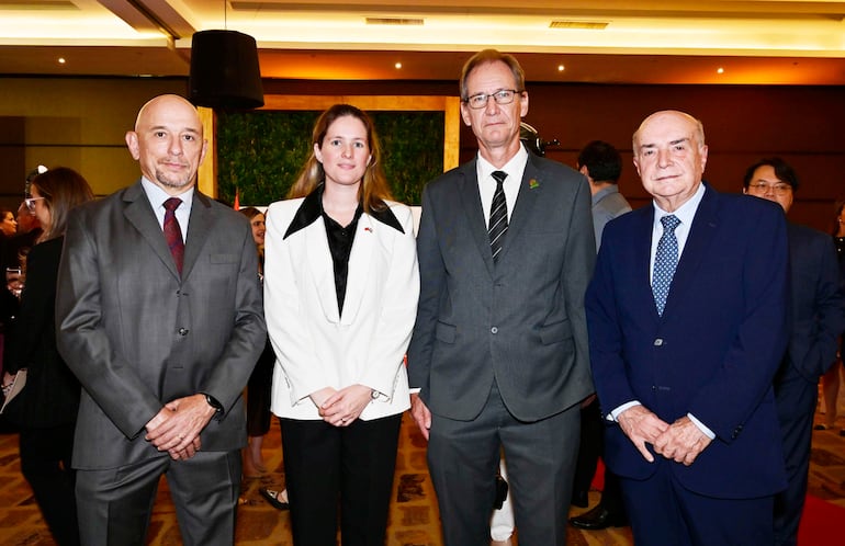 Cuatro personas conversan: hombre calvo en traje gris, mujer con blazer blanco, hombre con gafas en traje oscuro y hombre mayor en traje azul.