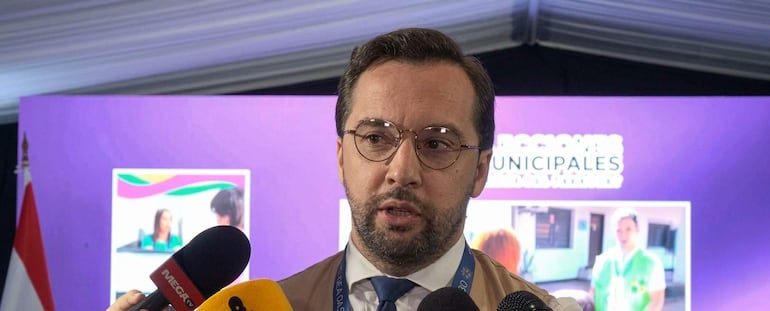 Hombre con cabello corto y barba habla en rueda de prensa, rodeado de micrófonos y fondo morado con imágenes relacionadas con salud y educación.
