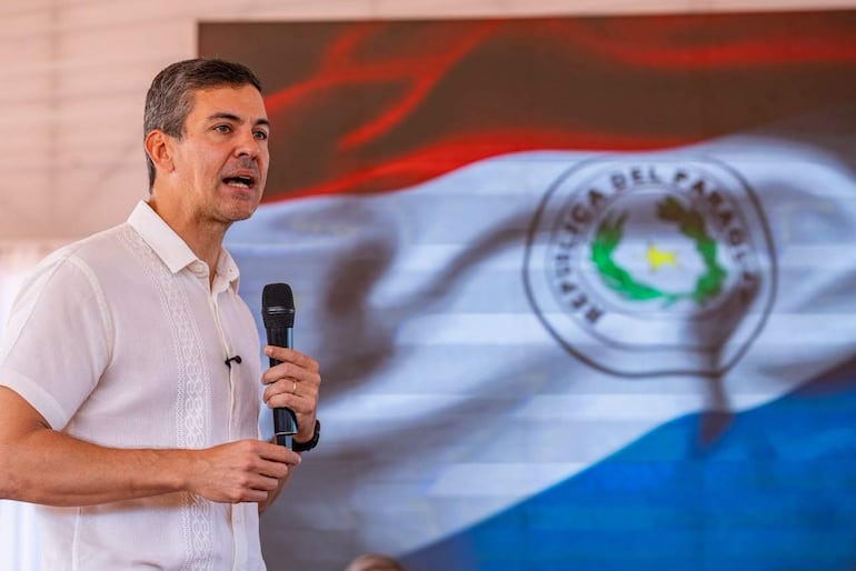 Santiago Peña de pie con camisa blanca y micrófono, hablando en un acto formal ante una pantalla con la bandera de Paraguay.
