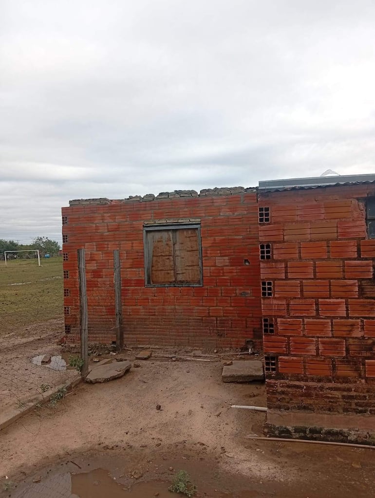 Edificio de ladrillo con ventanas cubiertas por tablas, terreno húmedo y campo de fútbol al fondo en un día nublado.