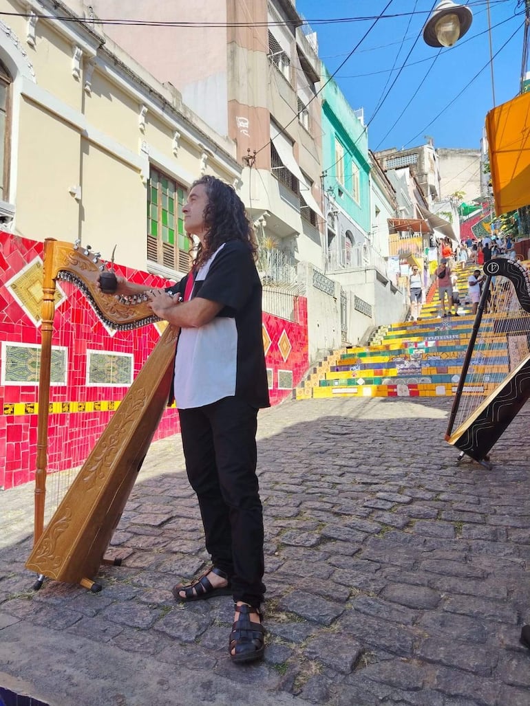 Hombre de cabello rizado tocando el arpa, vestido con camisa clara y pantalones oscuros, sosteniendo un mate, en un entorno vibrante.