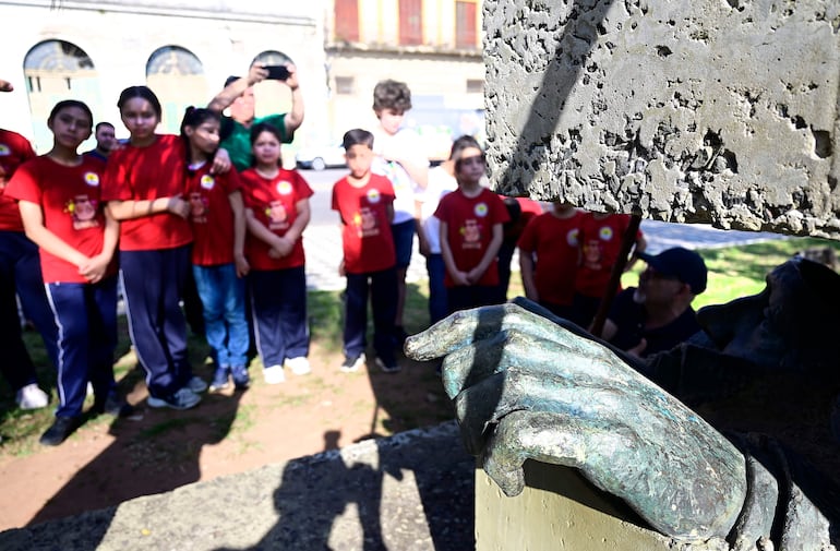 Grupo de diez niños con camisetas rojas, de pie en un semicírculo, miran un monumento en un ambiente con vegetación.