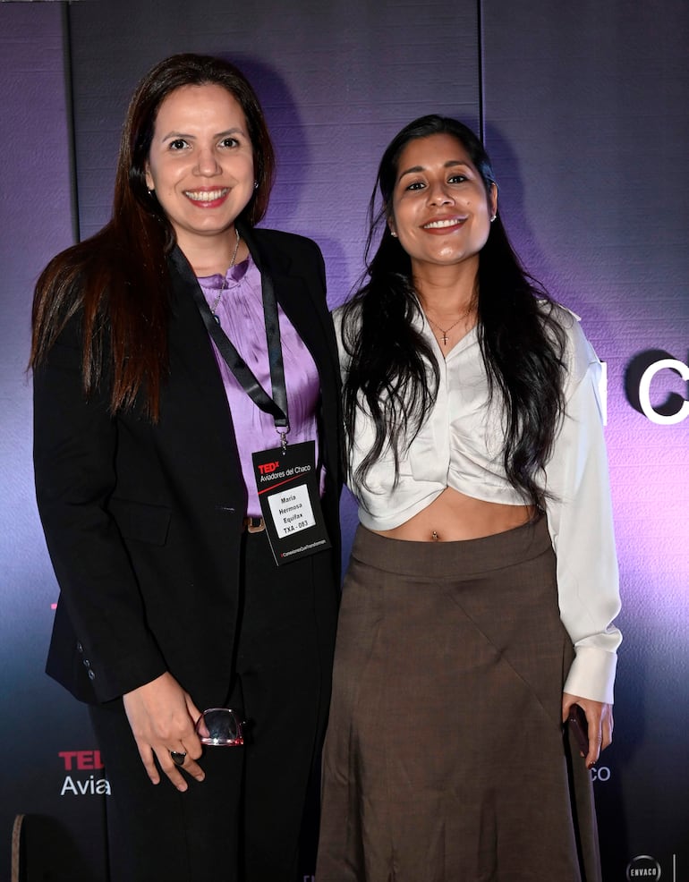 Dos mujeres, una con blusa negra y chaqueta, otra con camisa blanca, sonríen en un ambiente oscuro de evento formal.